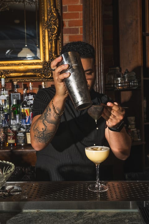 A bartender at The Alexander strains a shaken cocktail into a coupe on the bar, gilded mirror and back bar behind him