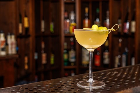 A dirty martini in a coupe with three green olives on a bamboo pick, resting on the brass bar rail