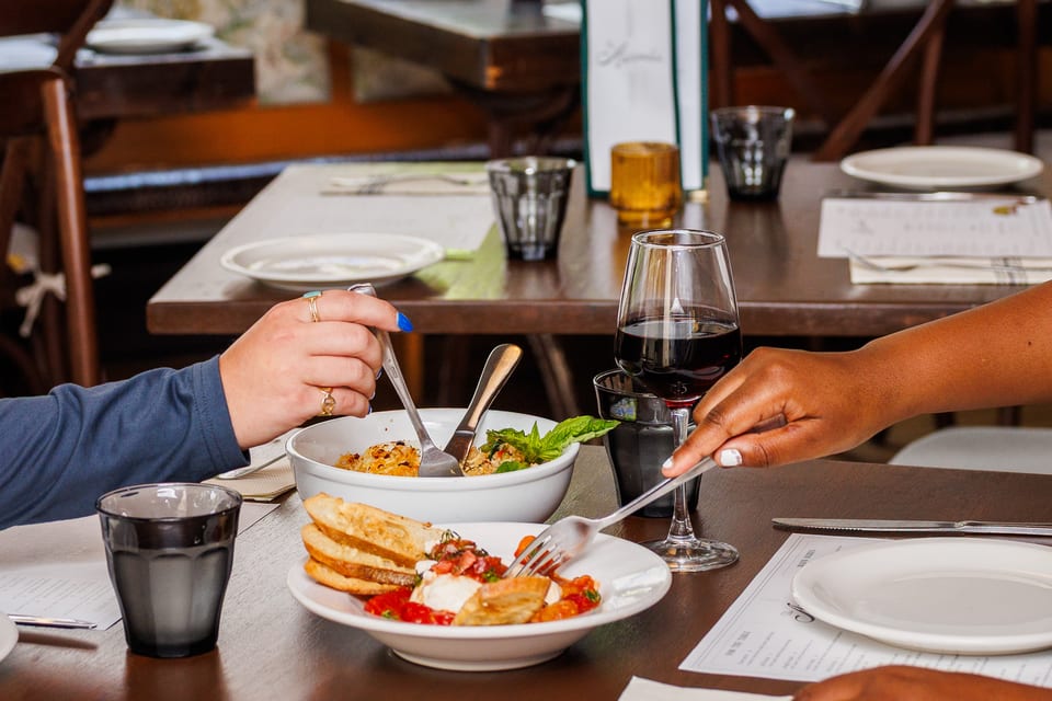 Two guests sharing bruschetta and red wine across a candlelit table — hands reaching mid-meal in a warm shared moment