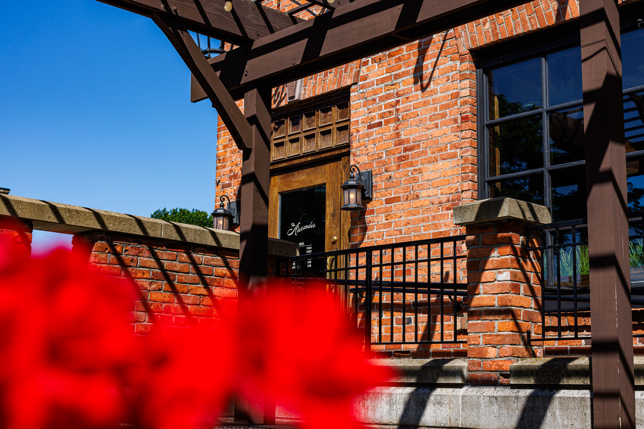 The Alexander entrance beneath the pergola — wooden door with the script logo, wrought-iron lanterns lit, and red flowers softly blurred in the foreground