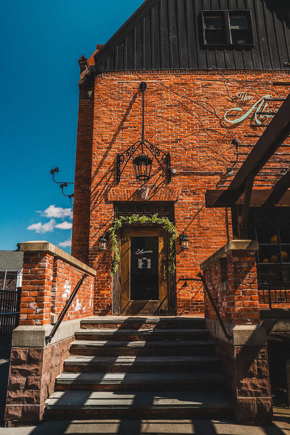 The Alexander entrance — wooden door framed by trailing greenery, ornate iron lantern, and brick stoop with stone steps leading up beneath the script signage