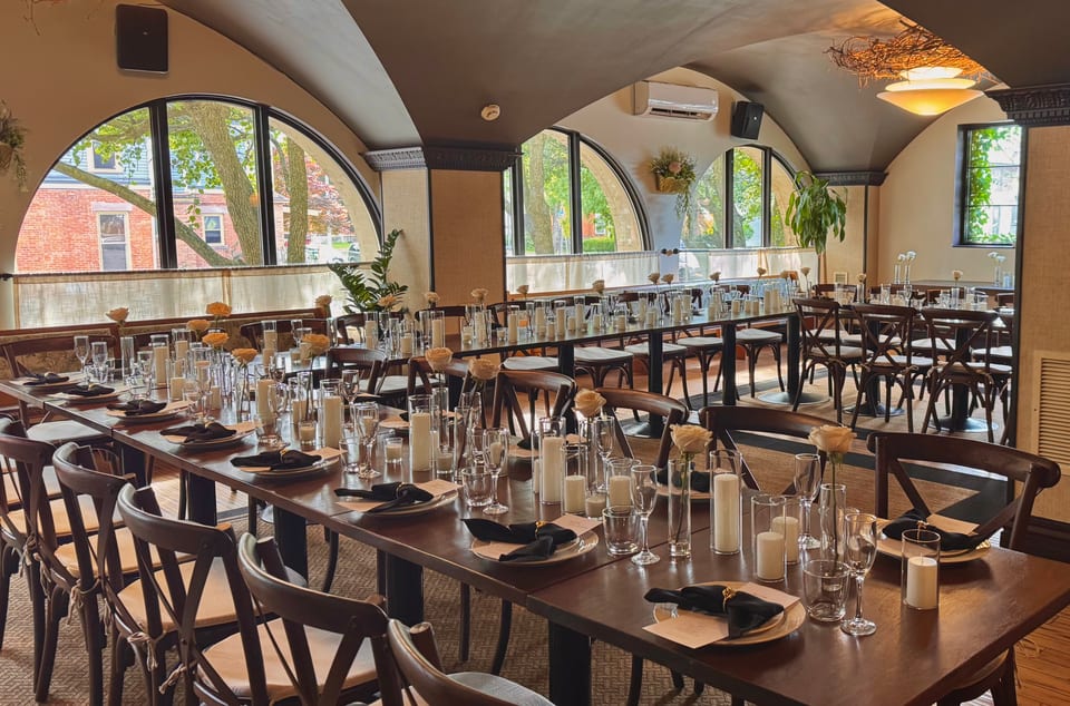 Banquet tables under arched windows set with candles, glassware, and greenery in daylight
