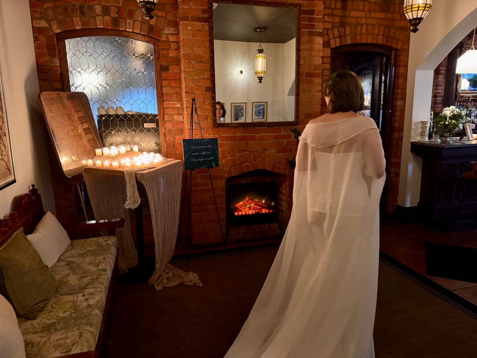 Bride in a long cape gazing into the fireplace, candles lit on the mantel, brick walls