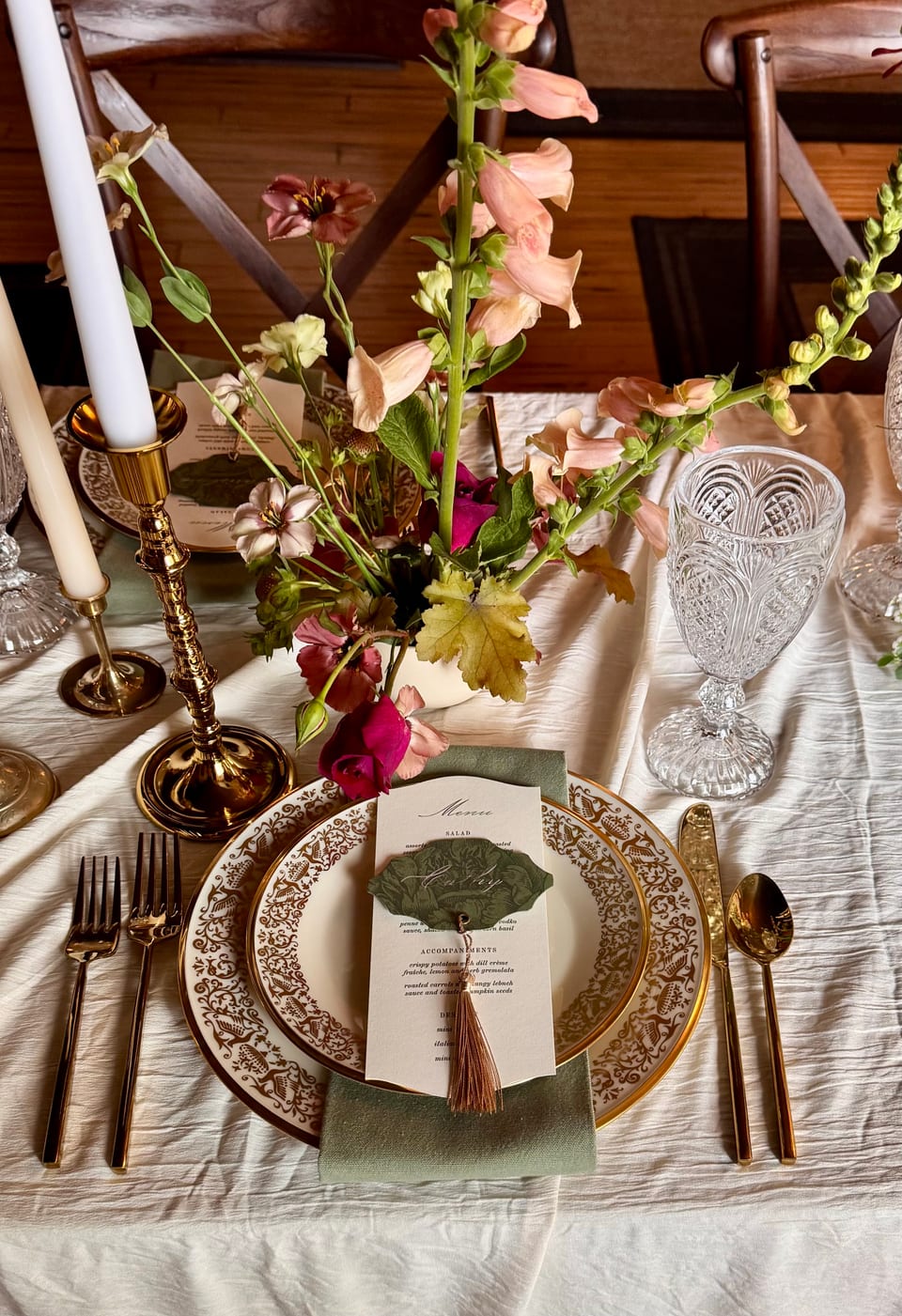 Place setting close-up — patterned china, gold flatware, sage napkin with tassel, snapdragon florals