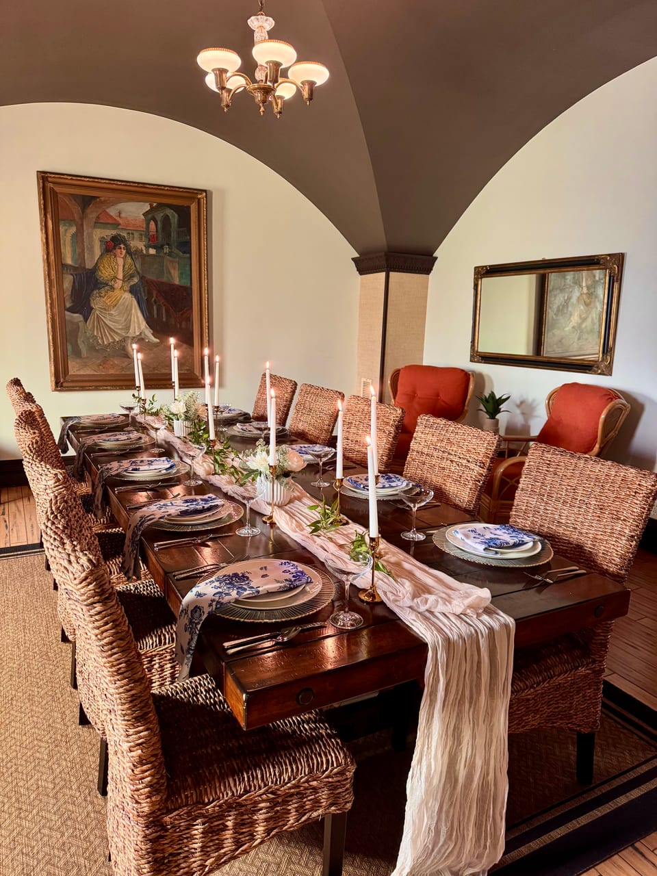 Long table set for an intimate dinner under vaulted ceilings — wicker chairs, candles, blue-and-white china, draped runner