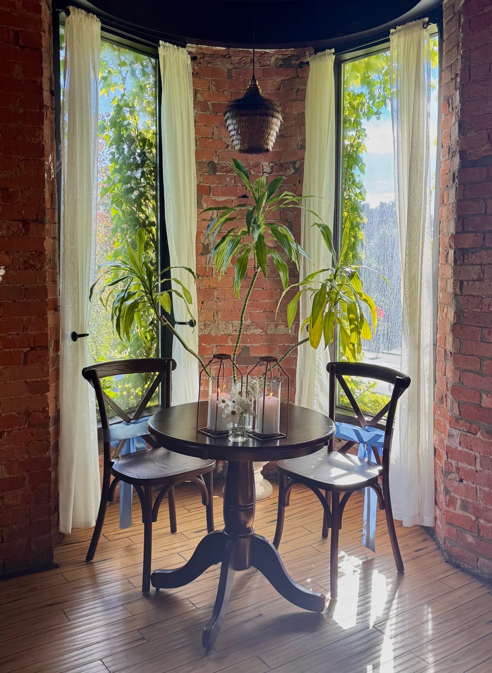 Round bistro table in the turret bay — potted greens, sheer curtains, brick walls in afternoon light