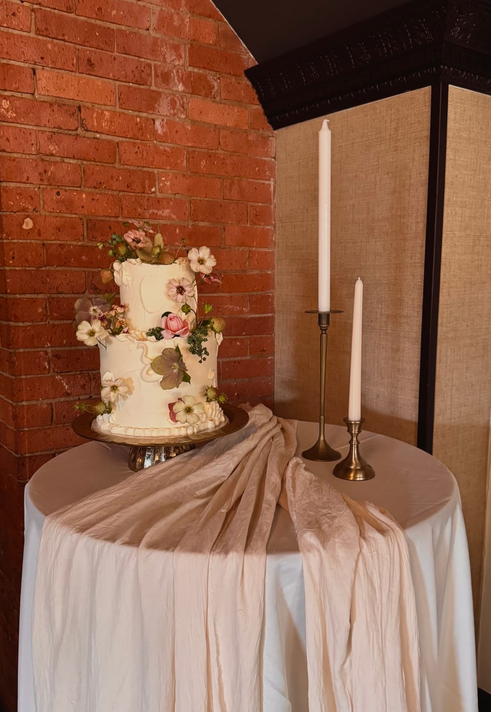 Two-tier wedding cake decorated with pressed flowers, draped linens and tall candles